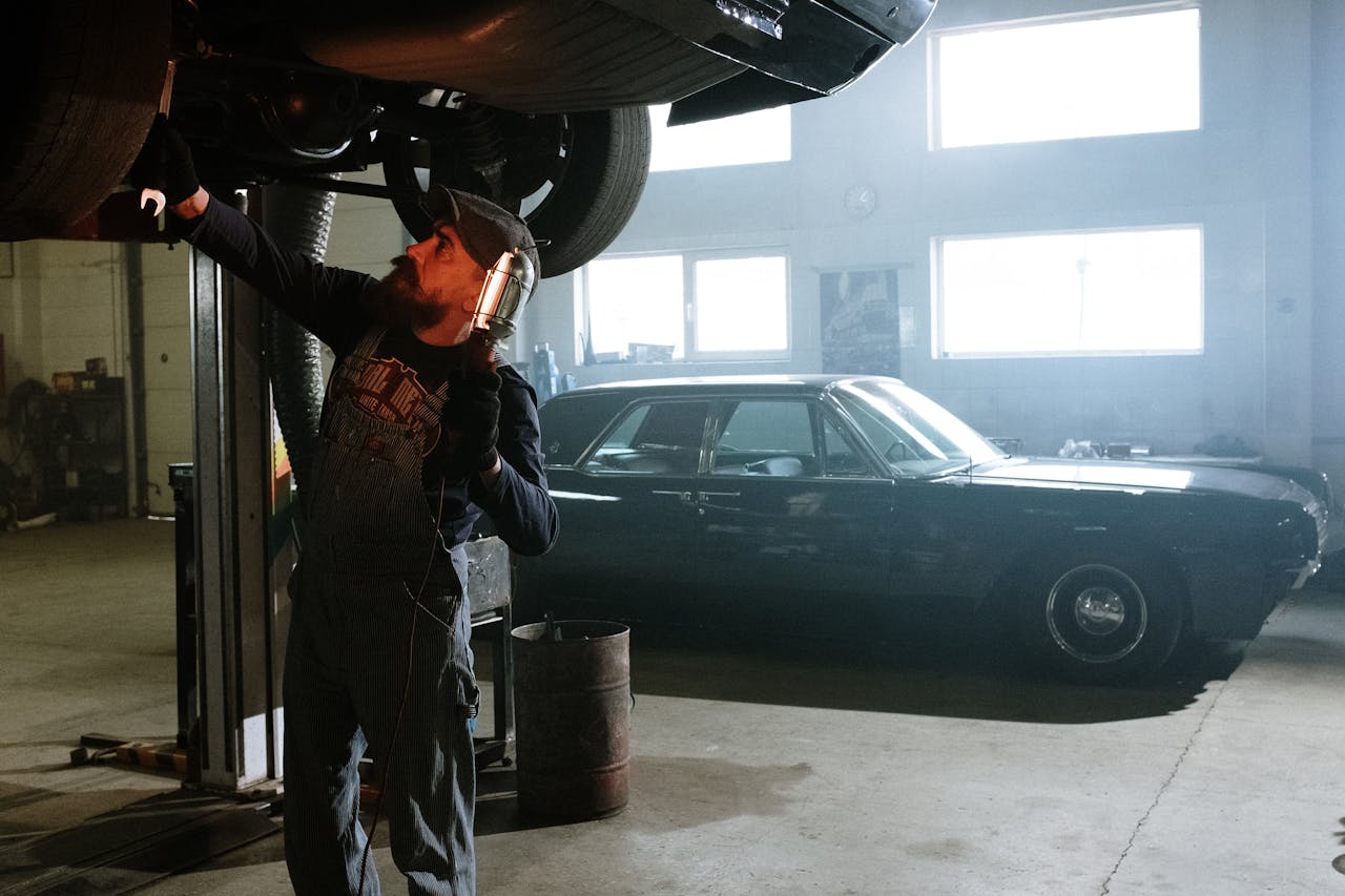 Mechanic working under a lifted car in an indoor garage setting with vintage vehicle in background.